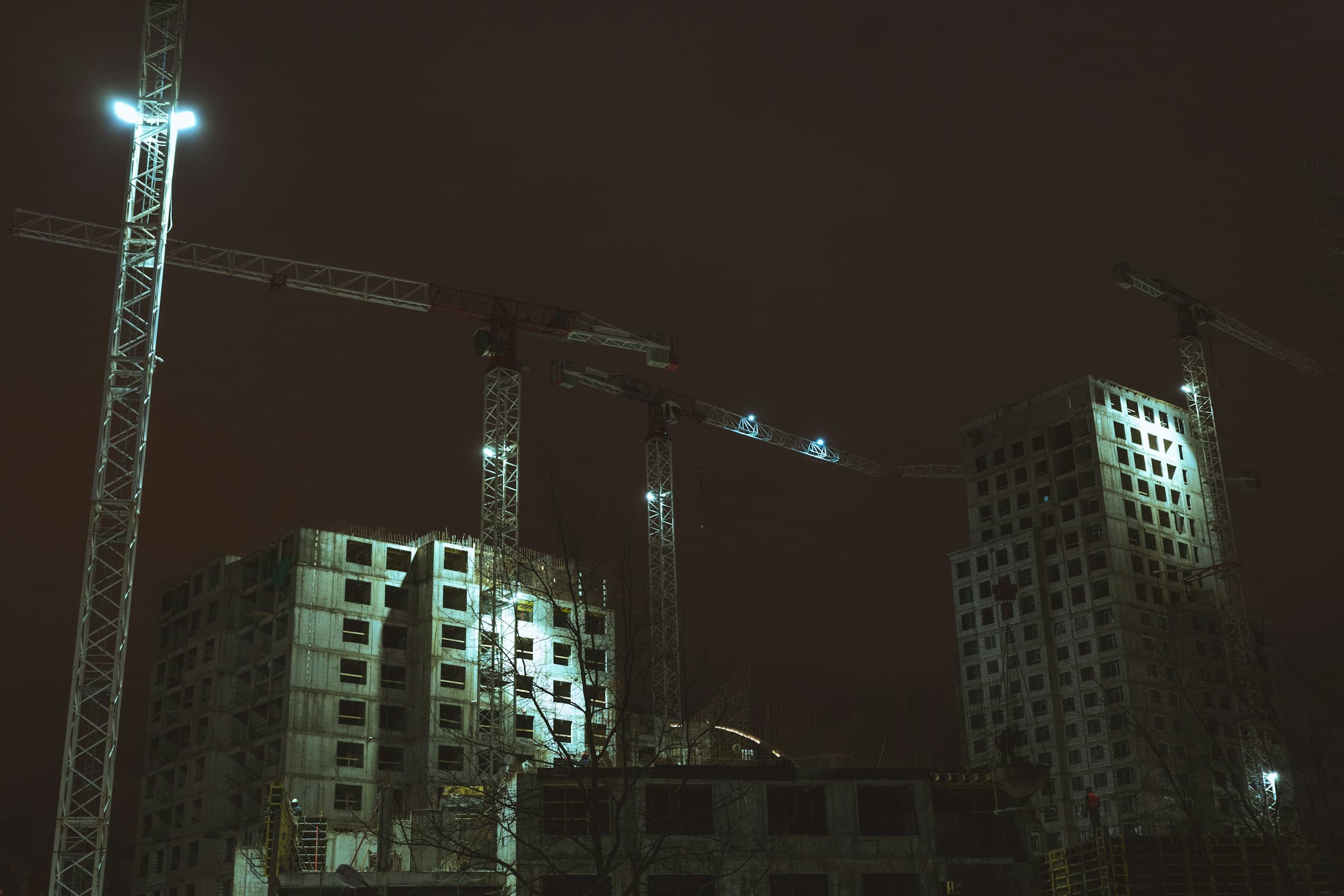 Construction site at night, tower cranes and floodlit concrete structures under a dark sky